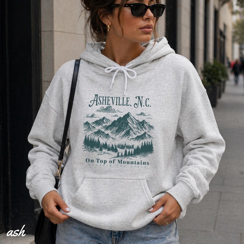 May include: Ash-colored hoodie with a front pocket and drawstring. The graphic on the front features the text "Asheville, N.C." above a mountain and forest scene, with the words "On Top of Mountains" below. The hoodie is a casual style.