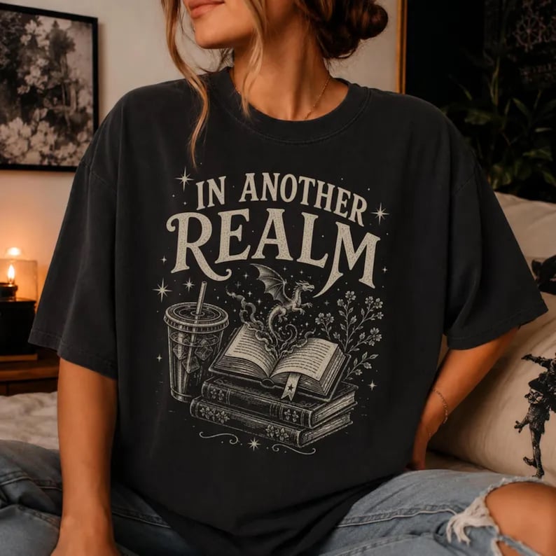 May include: A charcoal gray short-sleeve t-shirt featuring a graphic print with the text IN ANOTHER REALM. The design includes a stack of books, an open book with a small dragon emerging from the pages, a cold beverage cup, and star accents.