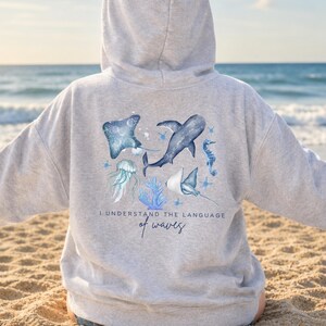 Pu&ograve; includere: Una felpa con cappuccio grigio chiaro con un design ad acquerello a tema marino. Il design include una manta, uno squalo, un cavalluccio marino e una medusa. Il testo "I understand the language of waves" &egrave; stampato sotto. La felpa con cappuccio &egrave; mostrata su una persona sulla spiaggia.
