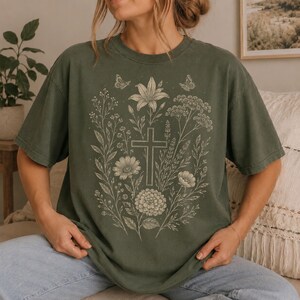 May include: A dark olive green short-sleeve t-shirt featuring a central graphic print in a light cream color. The design displays a cross surrounded by various botanical elements, including wildflowers, leaves, and two small butterflies.
