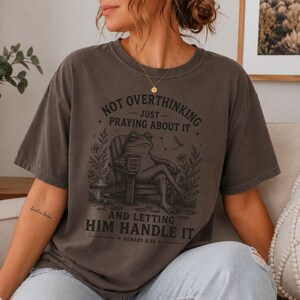 May include: A charcoal gray short-sleeve t-shirt featuring a graphic of a frog sitting in a wooden chair holding a mug. The text reads: NOT OVERTHINKING JUST PRAYING ABOUT IT AND LETTING HIM HANDLE IT ROMANS 8:28, surrounded by floral illustrations.