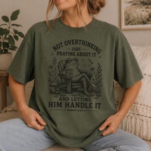 May include: An olive green short-sleeve t-shirt featuring a black graphic of a frog sitting in a chair holding a mug. The text reads: NOT OVERTHINKING JUST PRAYING ABOUT IT AND LETTING HIM HANDLE IT ROMANS 8:28. The design includes flowers and mushrooms.