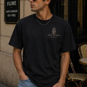 May include: A black short-sleeve t-shirt featuring a small graphic on the left chest. The graphic depicts a vintage lantern above the text "TRUST THE TRAIL" and "ISAIAH 41:10" in a light-colored font. The shirt has a crew neckline and a plain back.
