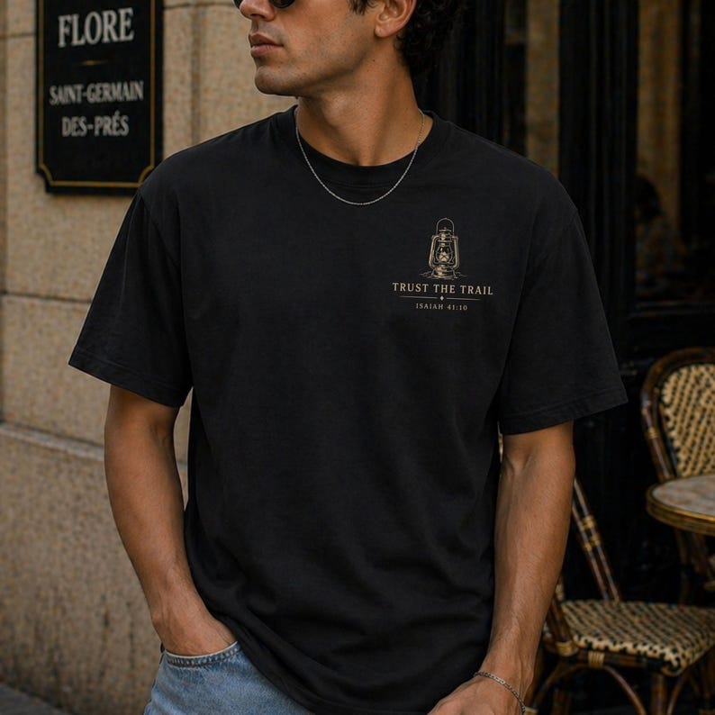 May include: A black short-sleeve t-shirt featuring a small graphic on the left chest. The graphic depicts a vintage lantern above the text "TRUST THE TRAIL" and "ISAIAH 41:10" in a light-colored font. The shirt has a crew neckline and a plain back.