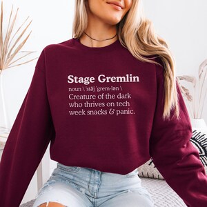 May include: A maroon sweatshirt with the text "Stage Gremlin" in white. The text is followed by a definition: "Creature of the dark who thrives on tech week snacks & panic."
