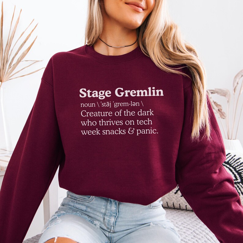 May include: A maroon sweatshirt with the text "Stage Gremlin" in white. The text is followed by a definition: "Creature of the dark who thrives on tech week snacks & panic."