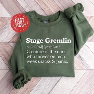 May include: An olive green sweatshirt with the text "Stage Gremlin" in white, along with a definition. A red "Fast Delivery" sticker is in the upper left corner. The sweatshirt has long sleeves.