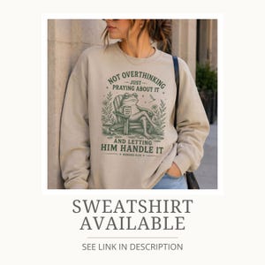 May include: A beige crewneck sweatshirt featuring a dark green graphic of a frog sitting in a wooden chair holding a mug. The text reads: NOT OVERTHINKING JUST PRAYING ABOUT IT AND LETTING HIM HANDLE IT, with the reference ROMANS 8:28 below.