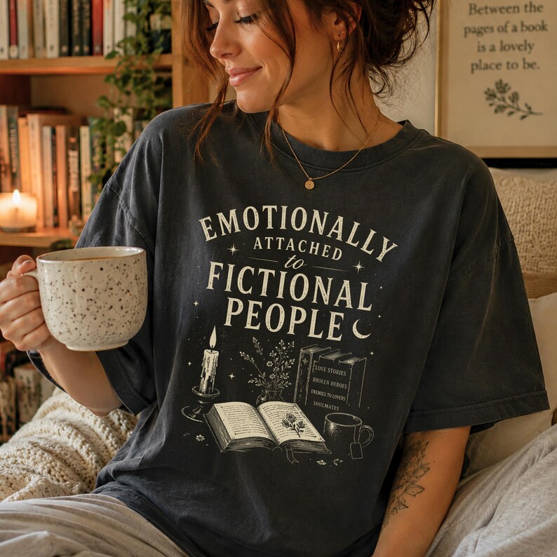 May include: Dark grey t-shirt with the text "EMOTIONALLY ATTACHED to FICTIONAL PEOPLE" in white. The graphic includes a candle, books, and a teacup. The person is holding a speckled ceramic mug.