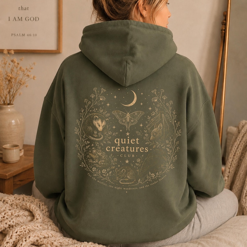 K&ouml;nnte beinhalten: Olivgr&uuml;ner Kapuzenpullover mit Kordelzug und gro&szlig;em Aufdruck auf dem R&uuml;cken. Das Design zeigt einen Mond, eine Motte, Sterne und die Worte "quiet cr&eacute;atures club" in einem kreisf&ouml;rmigen Design. Der Hoodie ist aus weichem Material.
