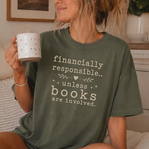 Pode incluir: Uma t-shirt de manga curta verde-azeitona com o texto em cor creme "financially responsible.. unless books are involved." acompanhado por um pequeno coração e estrelas. A t-shirt é combinada com uma caneca de cerâmica branca decorada com pequenos motivos florais castanhos.