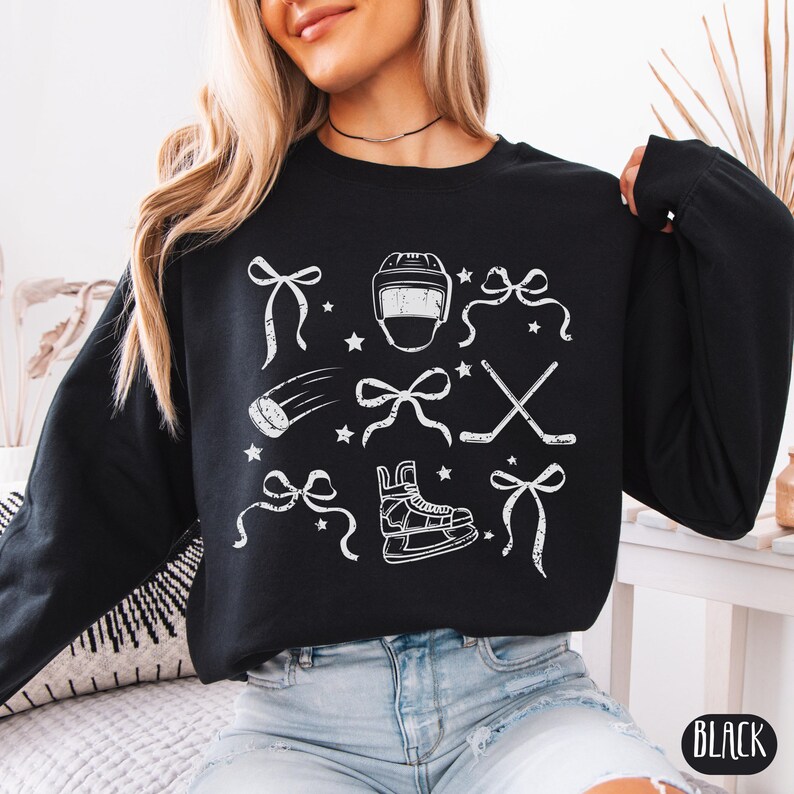 May include: Black sweatshirt with a white graphic design of hockey equipment and bows. The design includes a hockey helmet, hockey stick, hockey puck, and ice skates.