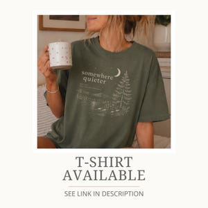 May include: Olive green t-shirt with a landscape graphic and the words "somewhere quieter". The design includes a crescent moon, a pine tree, and a lake scene. The shirt is a relaxed fit.