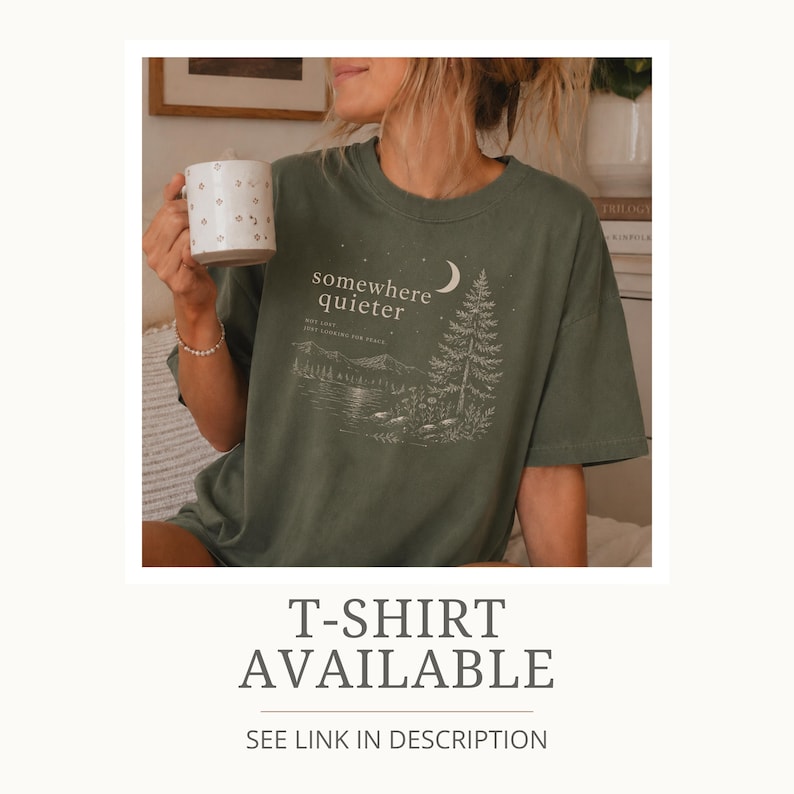 May include: Olive green t-shirt with a landscape graphic and the words "somewhere quieter". The design includes a crescent moon, a pine tree, and a lake scene. The shirt is a relaxed fit.