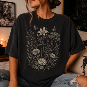 May include: A charcoal gray short-sleeve t-shirt featuring a central graphic print of a cross surrounded by detailed botanical illustrations of various flowers, leaves, and two small butterflies in a light cream color on the front chest.