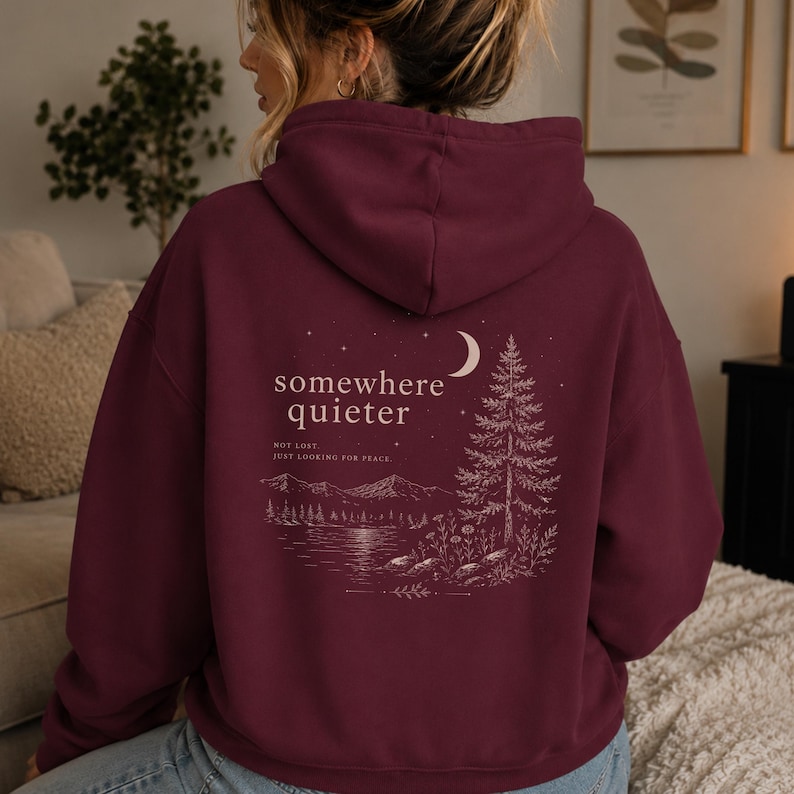 May include: Burgundy hoodie with a nature-themed graphic on the back. The design includes a crescent moon, a pine tree, mountains, and the text "somewhere quieter". The phrase "not lost, just looking for peace" is also included.