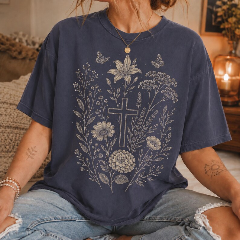 May include: A navy blue short-sleeve t-shirt featuring a central graphic print in a light cream color. The design displays a cross surrounded by various botanical elements, including flowers, leaves, and two small butterflies above the cross.