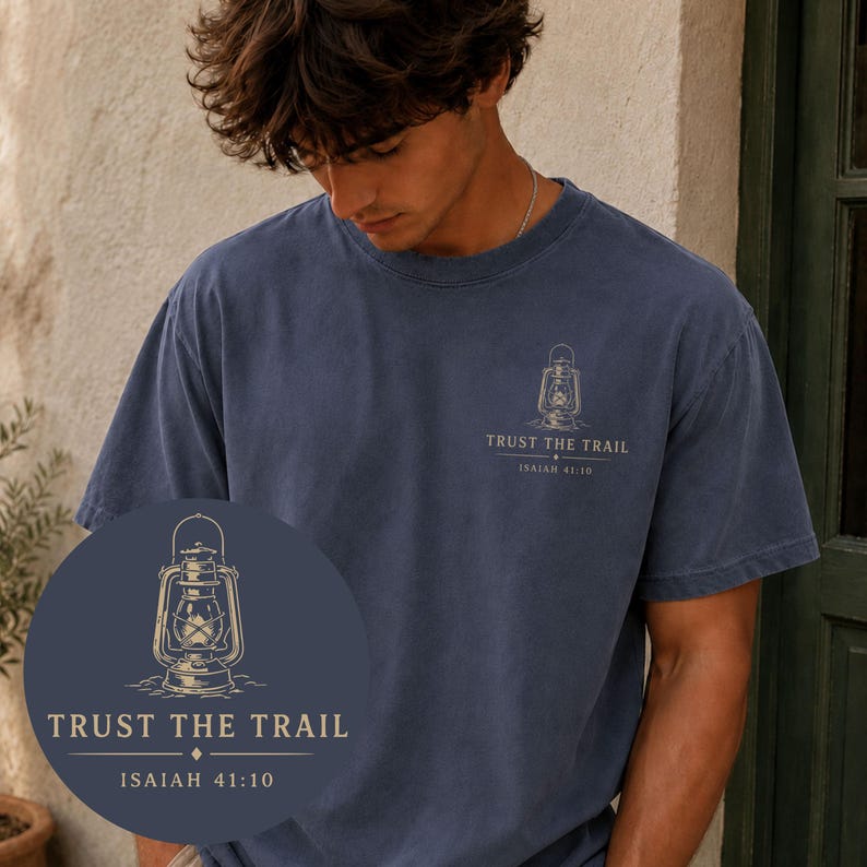 May include: A navy blue short-sleeve t-shirt featuring a graphic on the left chest. The graphic depicts a vintage lantern above the text "TRUST THE TRAIL" and "ISAIAH 41:10" in a light-colored font. The shirt has a crew neckline and a relaxed fit.