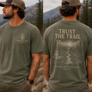 May include: An olive green t-shirt featuring a small lantern graphic and the text "TRUST THE TRAIL" on the front left chest. The back displays a large illustration of a forest landscape with a lantern, the text "TRUST THE TRAIL", and "FEAR NOT, FOR I AM WITH YOU. ISAIAH 41:10".