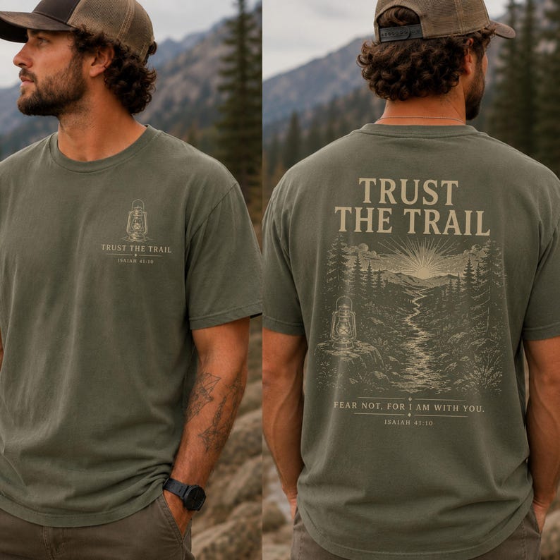 May include: An olive green t-shirt featuring a small lantern graphic and the text "TRUST THE TRAIL" on the front left chest. The back displays a large illustration of a forest landscape with a lantern, the text "TRUST THE TRAIL", and "FEAR NOT, FOR I AM WITH YOU. ISAIAH 41:10".