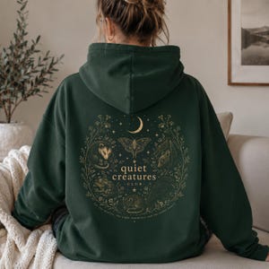 K&ouml;nnte beinhalten: Dunkelgr&uuml;ner Kapuzenpullover mit goldener Illustration auf dem R&uuml;cken. Die Illustration zeigt einen Mond, einen Schmetterling und verschiedene Tiere in einem Kreis. Der Text lautet "quiet creatures club".