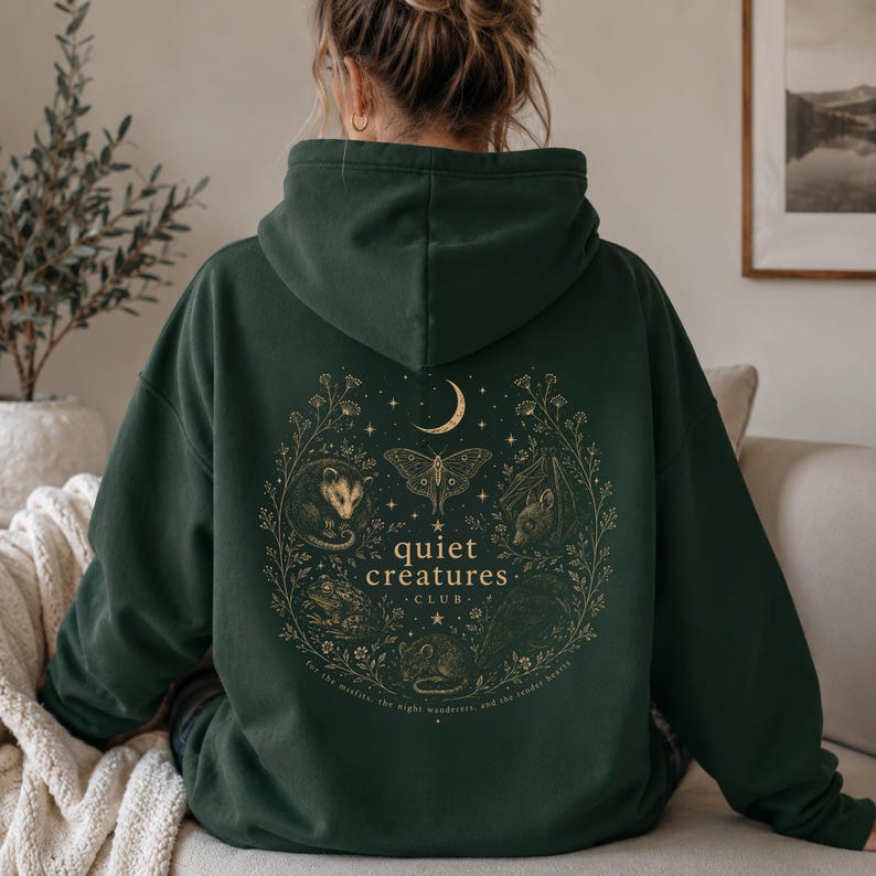 K&ouml;nnte beinhalten: Dunkelgr&uuml;ner Kapuzenpullover mit goldener Illustration auf dem R&uuml;cken. Die Illustration zeigt einen Mond, einen Schmetterling und verschiedene Tiere in einem Kreis. Der Text lautet "quiet creatures club".