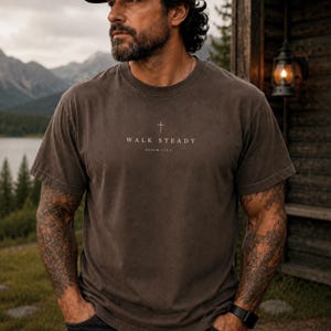 K&ouml;nnte beinhalten: Ein dunkelbraunes T-Shirt mit kurzen &Auml;rmeln und Rundhalsausschnitt, das ein kleines Kreuz-Symbol mittig &uuml;ber dem Text "WALK STEADY" und "PSALM 112:7" in einer hellen, neutralen Schrift auf der Brust zeigt.