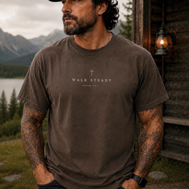 K&ouml;nnte beinhalten: Ein dunkelbraunes T-Shirt mit kurzen &Auml;rmeln und Rundhalsausschnitt, das ein kleines Kreuz-Symbol mittig &uuml;ber dem Text "WALK STEADY" und "PSALM 112:7" in einer hellen, neutralen Schrift auf der Brust zeigt.