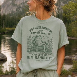 May include: A sage green short-sleeve t-shirt featuring a dark green graphic of a frog sitting in a chair holding a mug that says God's Timing Baby. The text reads Not Overthinking Just Praying About It And Letting Him Handle It Romans 8:28.