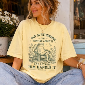 May include: A pale yellow short-sleeve t-shirt featuring a dark green graphic of a frog sitting in a wooden chair holding a mug that reads God's Timing Baby. The text reads Not Overthinking Just Praying About It And Letting Him Handle It Romans 8:28.