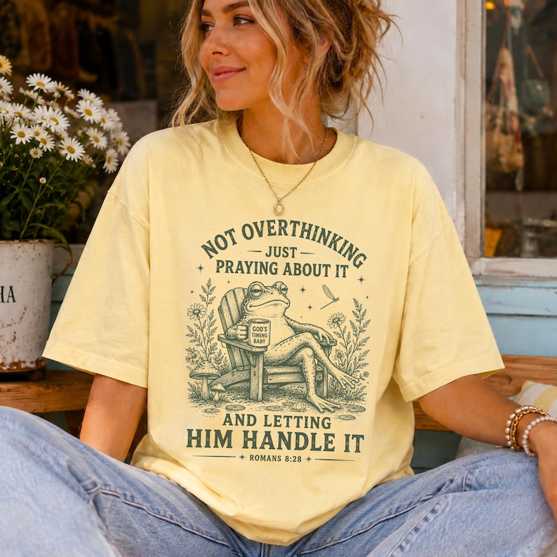 May include: A pale yellow short-sleeve t-shirt featuring a dark green graphic of a frog sitting in a wooden chair holding a mug that reads God's Timing Baby. The text reads Not Overthinking Just Praying About It And Letting Him Handle It Romans 8:28.