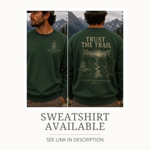 May include: A dark green crewneck sweatshirt featuring a small lantern graphic and the text "TRUST THE TRAIL" on the front left chest. The back displays a large illustration of a mountain landscape, a winding path, and a lantern with the text "TRUST THE TRAIL" and "FEAR NOT, FOR I AM WITH YOU. ISAIAH 41:10."