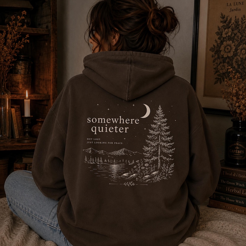 May include: A brown hoodie with the words "somewhere quieter" and a nature scene printed on the back. The design includes a crescent moon, a pine tree, mountains, and a lake. The text "NOT LOST JUST LOOKING FOR PEACE" is also printed on the back.