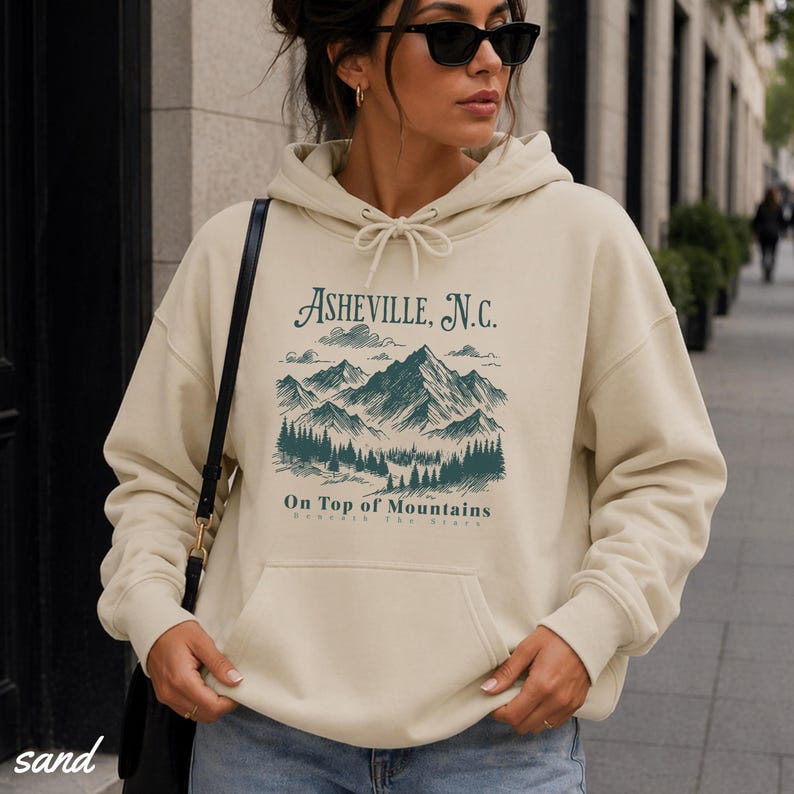 May include: Sand-colored hoodie with a drawstring hood and a front pocket. The front features a teal graphic of mountains and trees with the text "Asheville, N.C." and "On Top of Mountains". The word "sand" is printed at the bottom left.