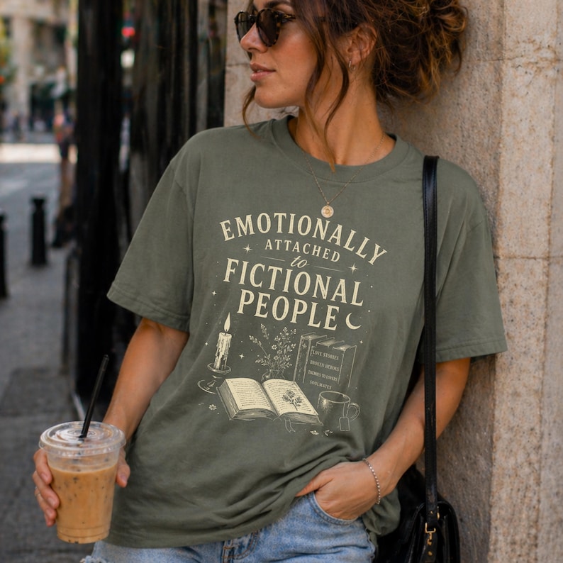 May include: An olive green t-shirt featuring the phrase "Emotionally Attached to Fictional People." The design includes a candle, an open book, and a cup. The shirt has a relaxed fit.