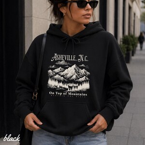 May include: Black hoodie with a drawstring hood and a front pocket. The graphic on the front features the text "Asheville, N.C." above a mountain scene with trees and the words "On Top of Mountains". The word "black" is printed at the bottom left.