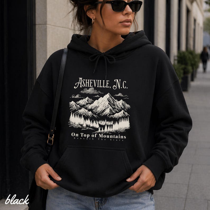 May include: Black hoodie with a drawstring hood and a front pocket. The graphic on the front features the text "Asheville, N.C." above a mountain scene with trees and the words "On Top of Mountains". The word "black" is printed at the bottom left.