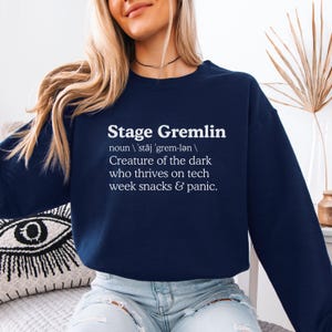 May include: Navy blue sweatshirt with the text "Stage Gremlin" in white, along with a definition. The definition describes a creature of the dark that thrives on tech week snacks and panic. A casual, comfortable top.