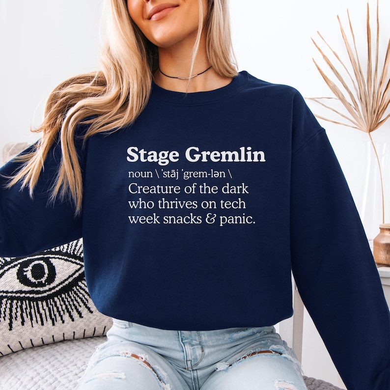 May include: Navy blue sweatshirt with the text "Stage Gremlin" in white, along with a definition. The definition describes a creature of the dark that thrives on tech week snacks and panic. A casual, comfortable top.