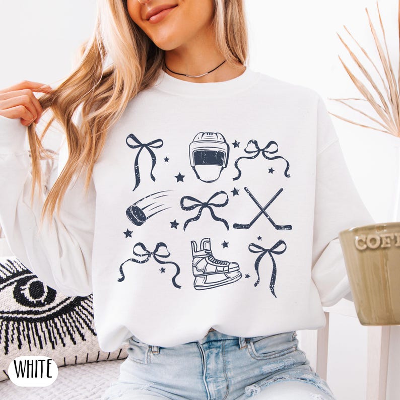May include: White sweatshirt with a blue graphic print of hockey equipment and bows. The graphic includes a hockey helmet, hockey stick, ice skates, and a puck.