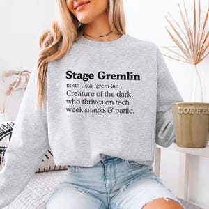 May include: A light gray sweatshirt with the text "Stage Gremlin" in black, along with a definition. The definition reads "Creature of the dark who thrives on tech week snacks & panic."