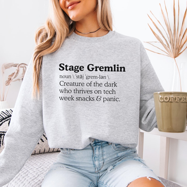 May include: A light gray sweatshirt with the text "Stage Gremlin" in black, along with a definition. The definition reads "Creature of the dark who thrives on tech week snacks & panic."