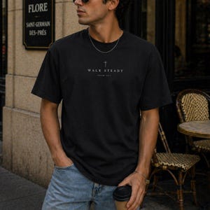 K&ouml;nnte beinhalten: Ein schwarzes kurz&auml;rmeliges T-Shirt mit einem kleinen wei&szlig;en Kreuz-Symbol, das mittig &uuml;ber dem Text "WALK STEADY" und "PSALM 119:5" in einer schlichten Schriftart auf der Brust gedruckt ist. Das Shirt hat einen klassischen Rundhalsausschnitt.