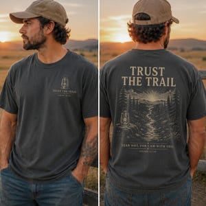 May include: A charcoal gray t-shirt featuring a small lantern graphic and the text "TRUST THE TRAIL" on the front left chest. The back displays a large illustration of a mountain trail, a lantern, and the text "TRUST THE TRAIL" and "FEAR NOT, FOR I AM WITH YOU. ISAIAH 41:10."
