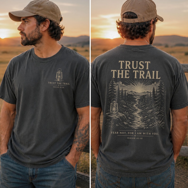May include: A charcoal gray t-shirt featuring a small lantern graphic and the text "TRUST THE TRAIL" on the front left chest. The back displays a large illustration of a mountain trail, a lantern, and the text "TRUST THE TRAIL" and "FEAR NOT, FOR I AM WITH YOU. ISAIAH 41:10."