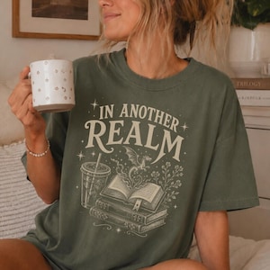 May include: An olive green t-shirt featuring a cream-colored graphic print with the text "IN ANOTHER REALM" above an illustration of an open book, a small dragon, a cup with a straw, and floral elements, all accented by small star symbols.