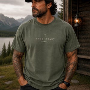 K&ouml;nnte beinhalten: Ein olivgr&uuml;nes Kurzarm-T-Shirt mit Rundhalsausschnitt, das ein kleines wei&szlig;es Kreuzsymbol &uuml;ber dem Text "WALK STEADY" und "PSALM 112:7" in einer klaren, zentrierten Schrift auf der Brustpartie des Kleidungsst&uuml;cks aufweist.