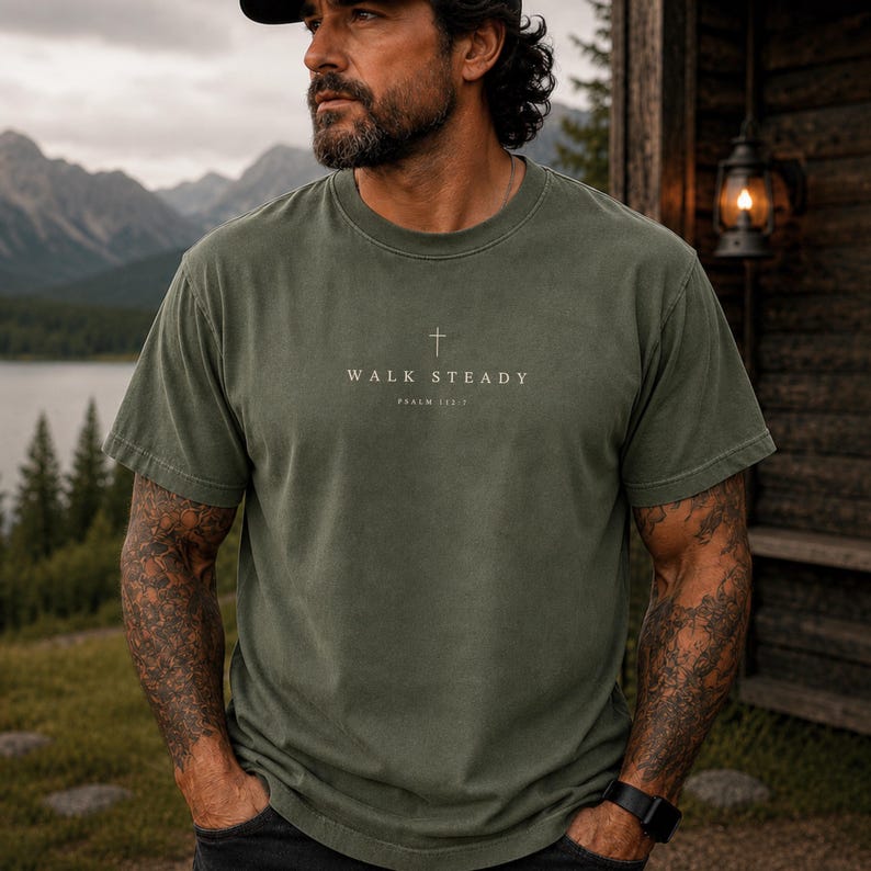 K&ouml;nnte beinhalten: Ein olivgr&uuml;nes Kurzarm-T-Shirt mit Rundhalsausschnitt, das ein kleines wei&szlig;es Kreuzsymbol &uuml;ber dem Text "WALK STEADY" und "PSALM 112:7" in einer klaren, zentrierten Schrift auf der Brustpartie des Kleidungsst&uuml;cks aufweist.