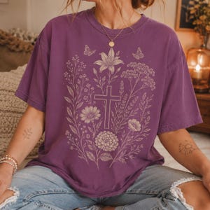 May include: A purple short-sleeve t-shirt featuring a cream-colored graphic print of a central cross surrounded by various wildflowers, leaves, and two small butterflies. The design is symmetrical and detailed, covering the center of the shirt.