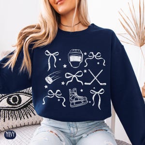May include: A navy blue sweatshirt with a white graphic design featuring hockey-themed illustrations, including a hockey helmet, skates, sticks, and bows.